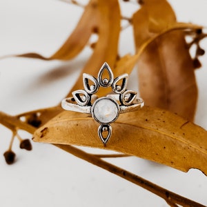 Puede incluir: Un anillo de plata con una piedra blanca en el centro y un diseño floral alrededor de la piedra. El anillo está sobre una hoja marrón.