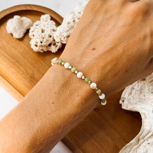 May include: A beaded bracelet featuring alternating round white beads, round sage green beads, and small metallic gold-toned cube spacers. The bracelet is strung on a flexible cord and sits on a wrist, showcasing a repeating pattern of these elements.