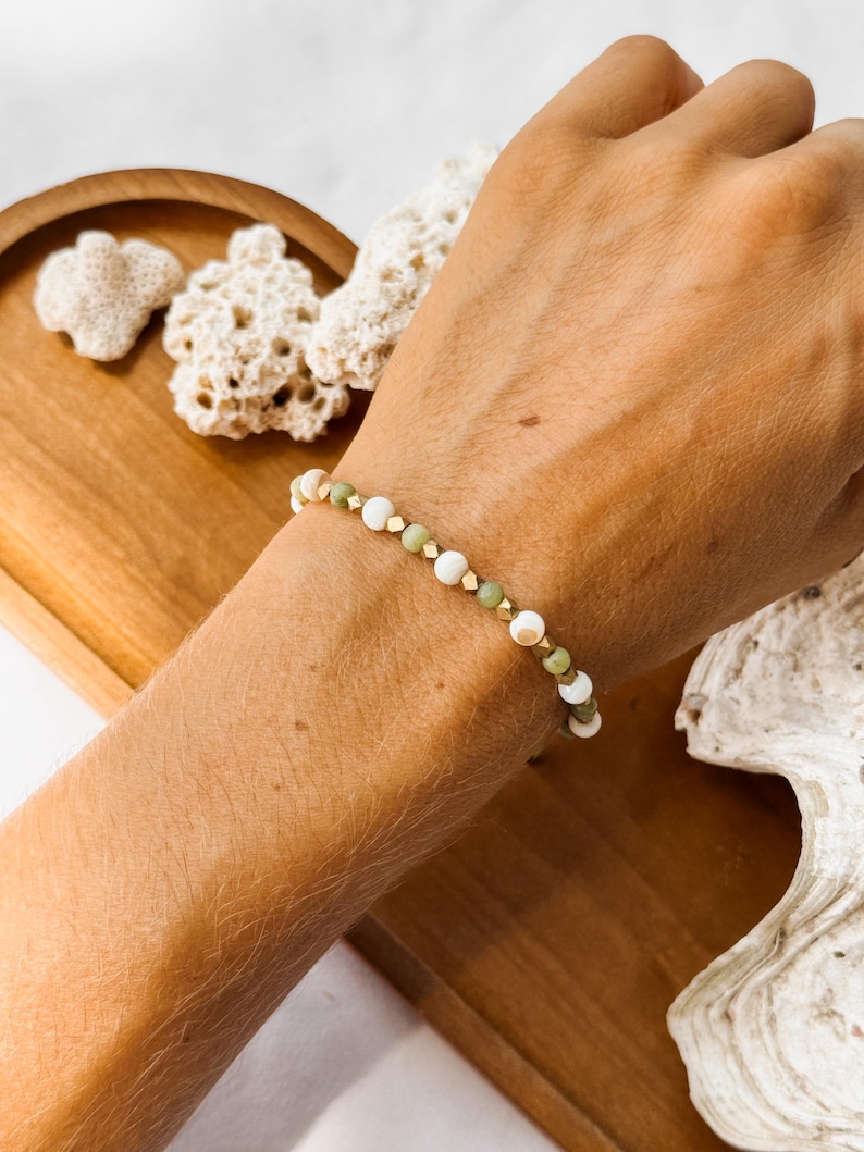 May include: A beaded bracelet featuring alternating round white beads, round sage green beads, and small metallic gold-toned cube spacers. The bracelet is strung on a flexible cord and sits on a wrist, showcasing a repeating pattern of these elements.