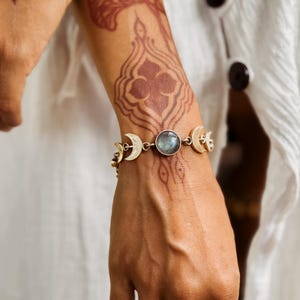 May include: A gold-toned bracelet featuring a central round iridescent gemstone flanked by crescent moon charms. The bracelet is constructed with metal links connecting the moon motifs and the circular stone, creating a celestial-inspired design.