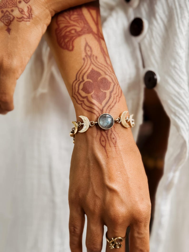 May include: A gold-toned bracelet featuring a central round iridescent gemstone flanked by crescent moon charms. The bracelet is constructed with metal links connecting the moon motifs and the circular stone, creating a celestial-inspired design.