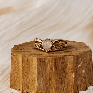Puede incluir: Un anillo de oro con una piedra preciosa blanca en forma de pera engastada en un diseño delicado y ornamentado. El anillo se muestra en un soporte de madera.