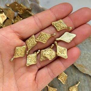 Pode incluir: Uma cole&ccedil;&atilde;o de pequenos pingentes dourados em forma de diamante. Cada pingente apresenta um design em relevo, possivelmente uma serpente estilizada, e um pequeno la&ccedil;o para fixa&ccedil;&atilde;o. Os pingentes s&atilde;o exibidos em uma m&atilde;o, sugerindo que s&atilde;o componentes de joias ou amuletos.