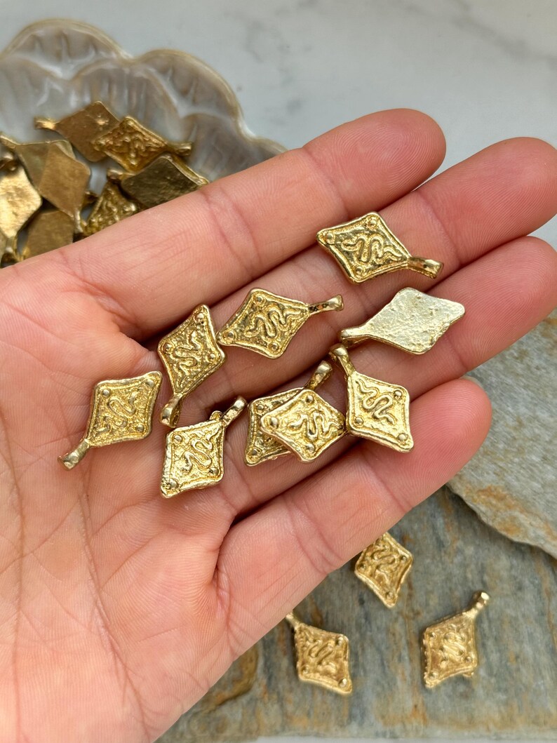 Pode incluir: Uma cole&ccedil;&atilde;o de pequenos pingentes dourados em forma de diamante. Cada pingente apresenta um design em relevo, possivelmente uma serpente estilizada, e um pequeno la&ccedil;o para fixa&ccedil;&atilde;o. Os pingentes s&atilde;o exibidos em uma m&atilde;o, sugerindo que s&atilde;o componentes de joias ou amuletos.