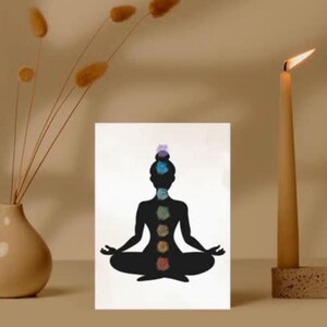 Może przedstawiać: Biały, prostokątny nadruk przedstawiający czarną sylwetkę osoby w pozycji siedzącej jogi, z kolorowymi symbolami czakr ułożonymi wzdłuż kręgosłupa. W tle zapalona świeca i wazon z suszonymi kwiatami.