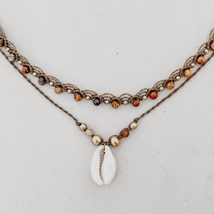 May include: A layered necklace with brown cord and brown and gold beads. The bottom layer has a white seashell pendant.