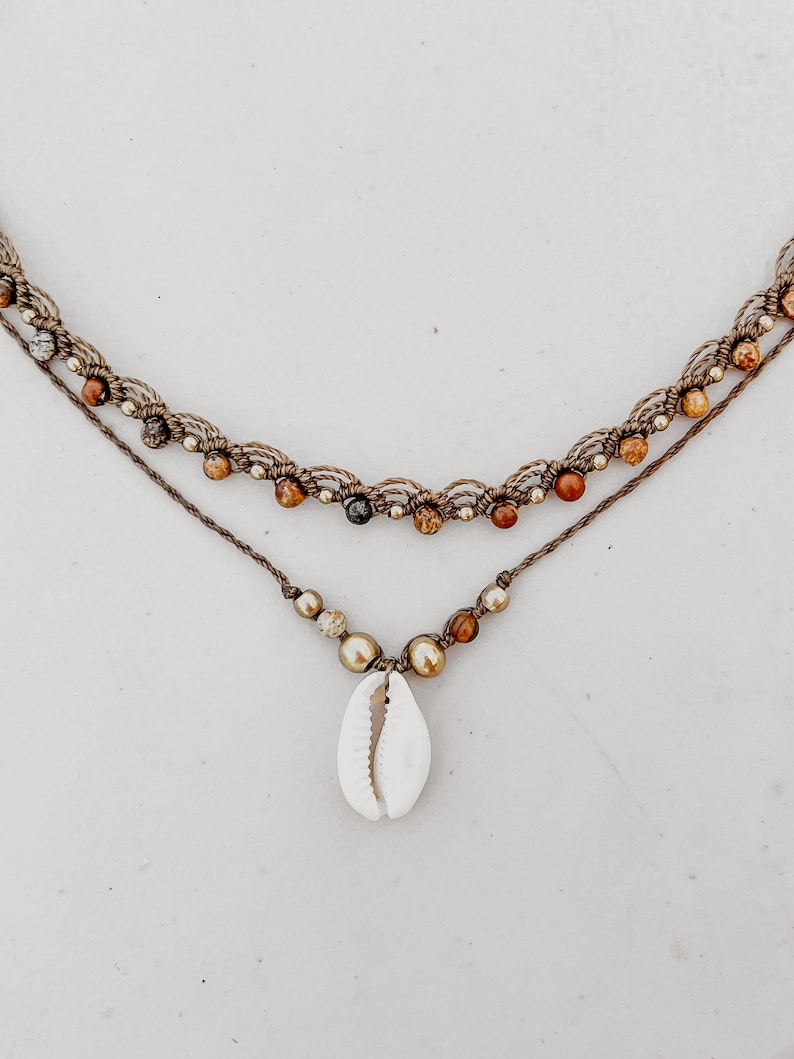 May include: A layered necklace with brown cord and brown and gold beads. The bottom layer has a white seashell pendant.