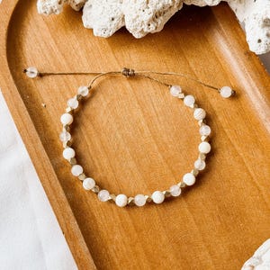 May include: A beaded bracelet featuring alternating round white beads, translucent clear beads, and small metallic gold-toned geometric spacers. The bracelet is constructed with a thin brown cord and includes an adjustable sliding knot closure.