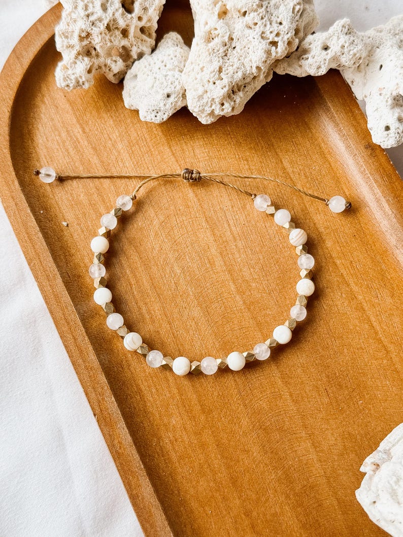 May include: A beaded bracelet featuring alternating round white beads, translucent clear beads, and small metallic gold-toned geometric spacers. The bracelet is constructed with a thin brown cord and includes an adjustable sliding knot closure.