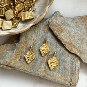 Puede incluir: Tres abalorios de metal dorado con un dise&ntilde;o geom&eacute;trico. Los abalorios son de forma cuadrada con un patr&oacute;n intrincado en relieve. Se muestran sobre una piedra marr&oacute;n.