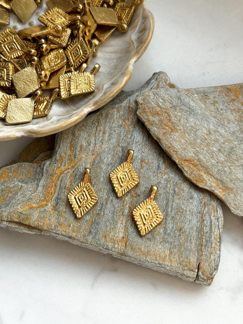 Puede incluir: Tres abalorios de metal dorado con un dise&ntilde;o geom&eacute;trico. Los abalorios son de forma cuadrada con un patr&oacute;n intrincado en relieve. Se muestran sobre una piedra marr&oacute;n.