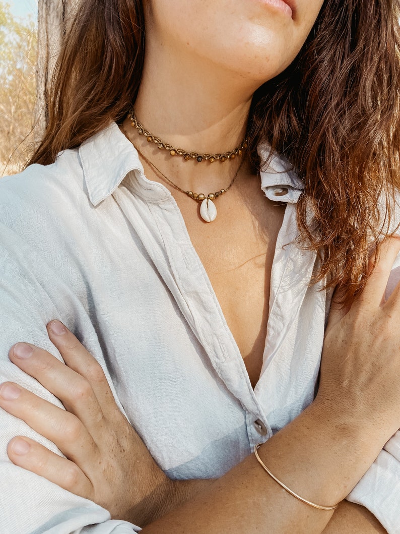 May include: A layered necklace with a shell pendant. The top layer is a beaded chain with gold-toned beads. The bottom layer is a simple cord with gold-toned beads. The necklace is worn by a person with long brown hair.