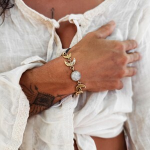 May include: A gold-toned metal bracelet featuring a series of crescent moon charms and a central round, translucent white stone. The bracelet is constructed with linked metal segments that alternate between the moon motifs and the circular stone.