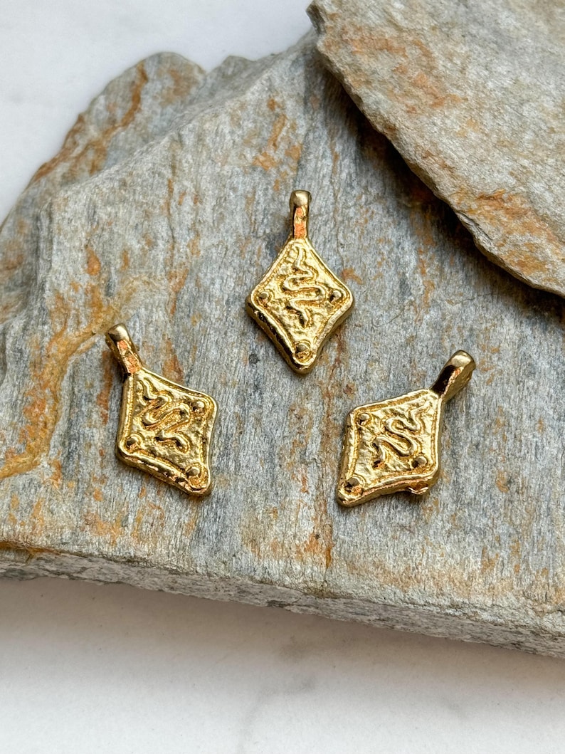 Pode incluir: Tr&ecirc;s pingentes dourados em forma de diamante com um design de cobra texturizado. Cada pingente tem uma pequena argola na parte superior para pendurar. Os pingentes s&atilde;o exibidos em uma superf&iacute;cie de pedra cinza.