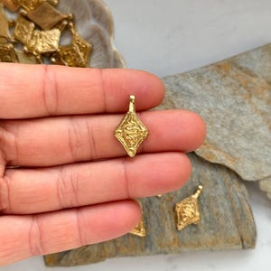 Pode incluir: Close-up de v&aacute;rios pingentes pequenos dourados em forma de losango. Um pingente &eacute; segurado entre os dedos, mostrando um design em relevo detalhado. Outros pingentes est&atilde;o espalhados em uma superf&iacute;cie de pedra e em um prato decorativo.