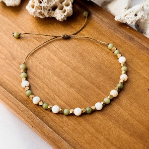 May include: A beaded bracelet featuring alternating round green stone beads, smooth white beads, and small metallic gold-toned faceted spacers. The beads are strung on a thin brown cord with an adjustable sliding knot closure and two small green beads at the ends.