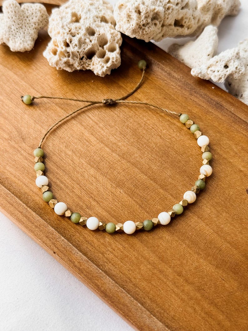 May include: A beaded bracelet featuring alternating round green stone beads, smooth white beads, and small metallic gold-toned faceted spacers. The beads are strung on a thin brown cord with an adjustable sliding knot closure and two small green beads at the ends.