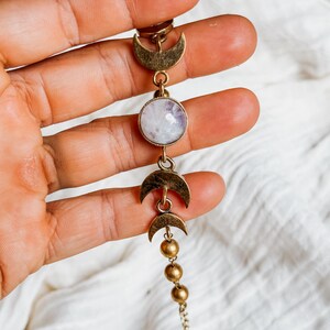 May include: A bracelet featuring a central round iridescent stone set in a brass-toned metal frame, flanked by two crescent moon charms and three small spherical beads, connected by a delicate chain link structure in a metallic finish.