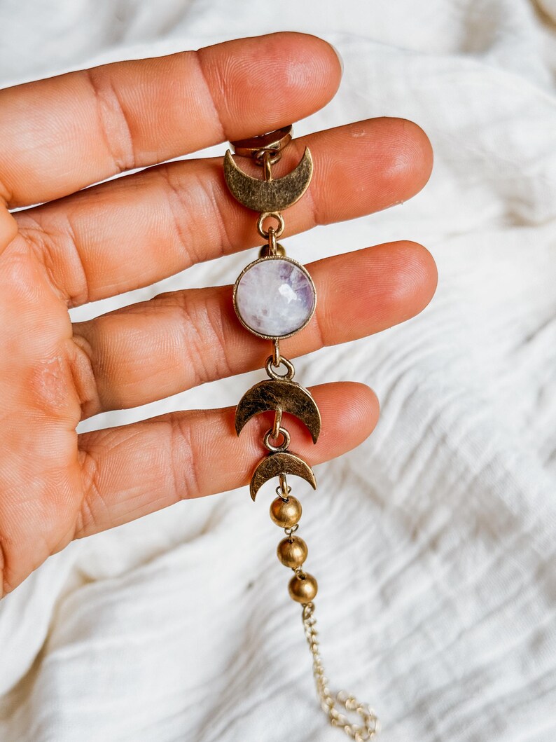 May include: A bracelet featuring a central round iridescent stone set in a brass-toned metal frame, flanked by two crescent moon charms and three small spherical beads, connected by a delicate chain link structure in a metallic finish.