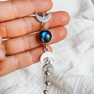 May include: A silver-toned bracelet featuring a central round labradorite gemstone with blue iridescence, flanked by two crescent moon charms and two smaller crescent moon charms, followed by a series of small silver-toned metal beads on a chain.