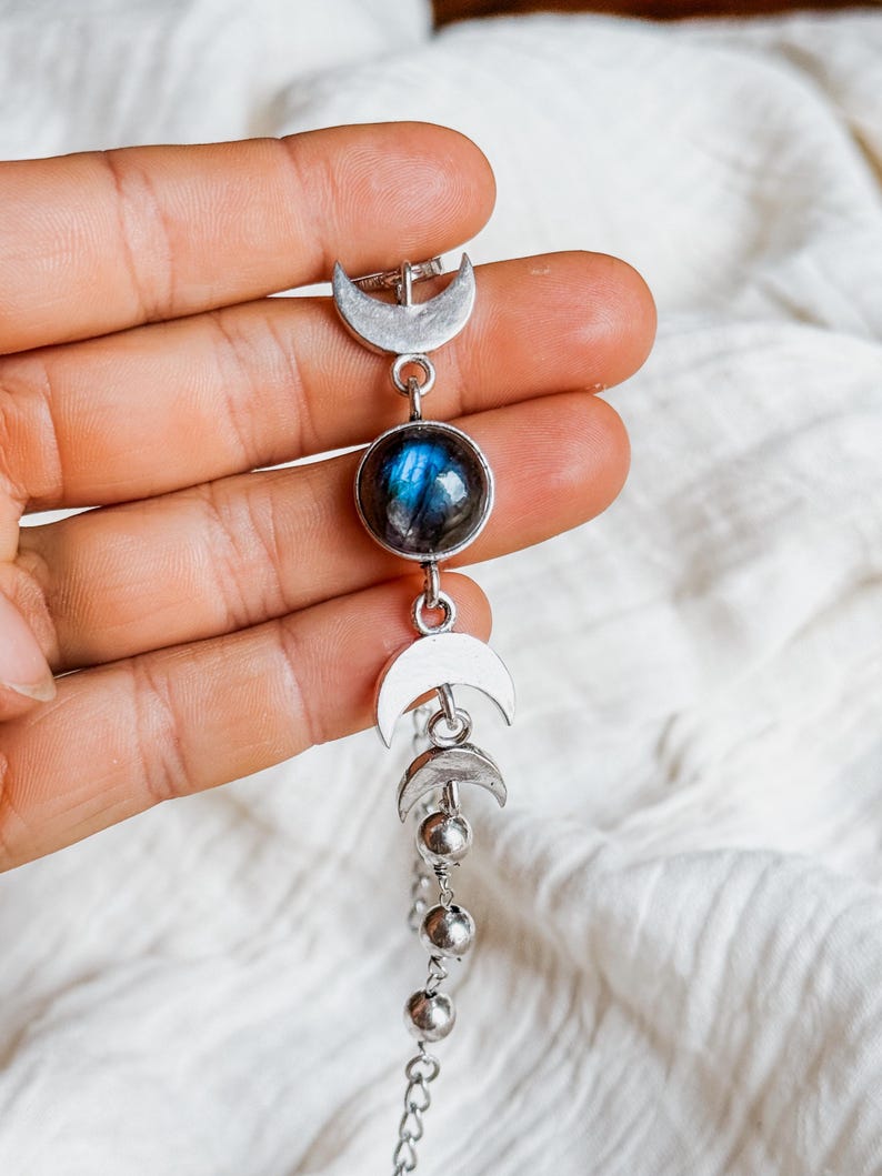 May include: A silver-toned bracelet featuring a central round labradorite gemstone with blue iridescence, flanked by two crescent moon charms and two smaller crescent moon charms, followed by a series of small silver-toned metal beads on a chain.
