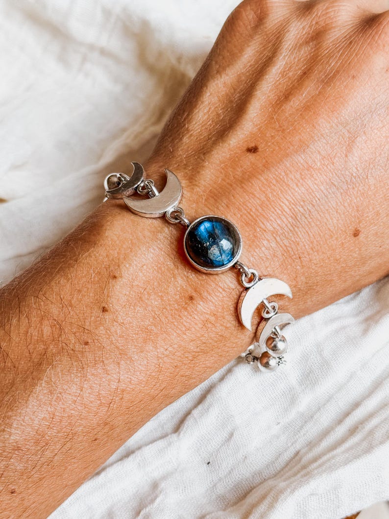 May include: A silver-toned bracelet featuring a central round cabochon stone with blue iridescent flashes, flanked by multiple crescent moon-shaped metal charms and small spherical beads connected by silver links in a chain design.