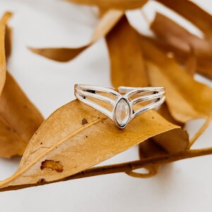 Puede incluir: Un anillo de plata con una piedra preciosa transparente y un diseño en forma de V. El anillo está sobre una hoja marrón.
