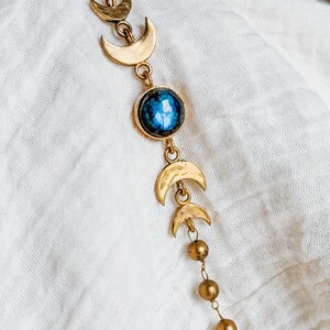 May include: A gold-toned chain necklace featuring a series of small crescent moon charms and spherical beads. The centerpiece is a round, polished blue gemstone with a mottled texture, set in a gold-toned metal frame, connected by delicate links.
