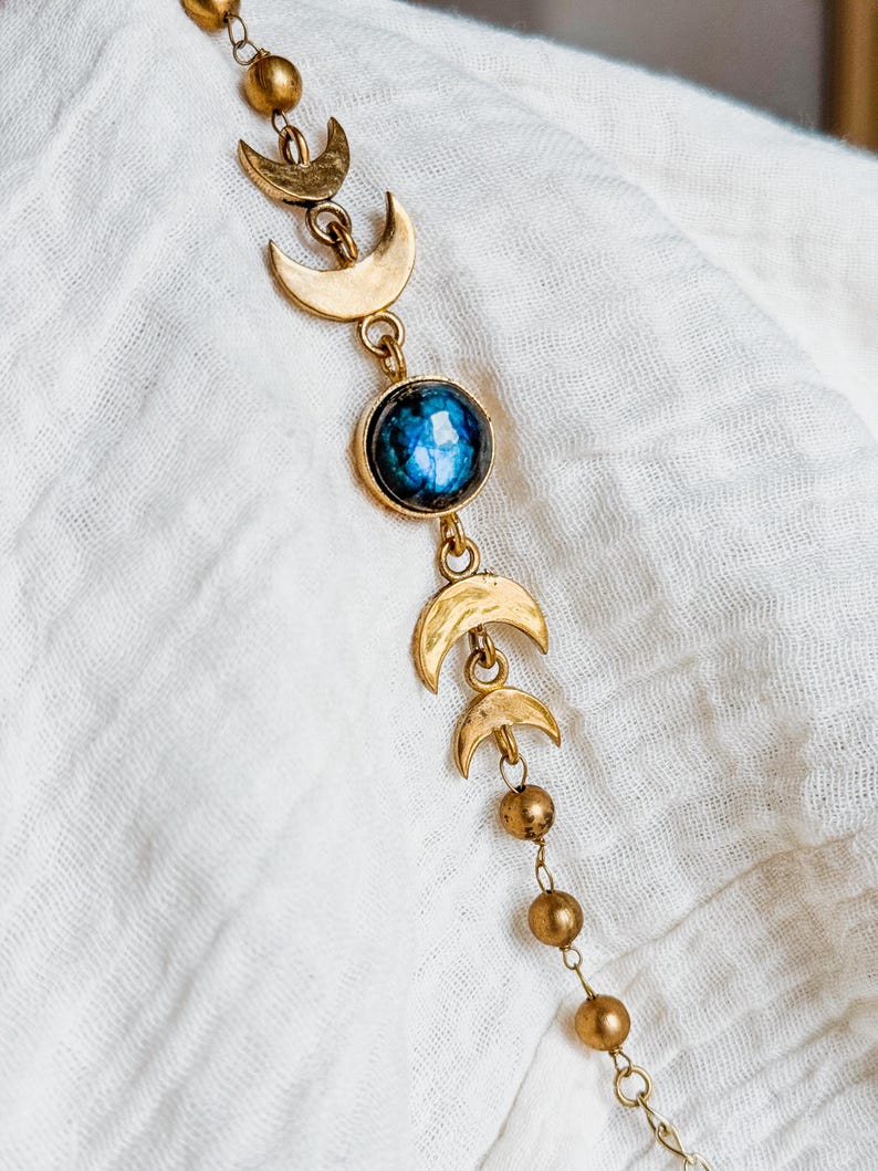 May include: A gold-toned chain necklace featuring a series of small crescent moon charms and spherical beads. The centerpiece is a round, polished blue gemstone with a mottled texture, set in a gold-toned metal frame, connected by delicate links.