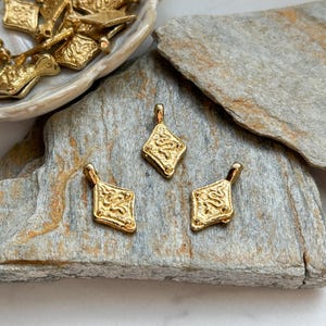 Pode incluir: Pingentes de metal dourado em forma de diamante com design ornamentado. Tr&ecirc;s pingentes s&atilde;o dispostos sobre uma superf&iacute;cie de pedra cinza, com pingentes adicionais em um prato decorativo. Os pingentes t&ecirc;m uma argola na parte superior.
