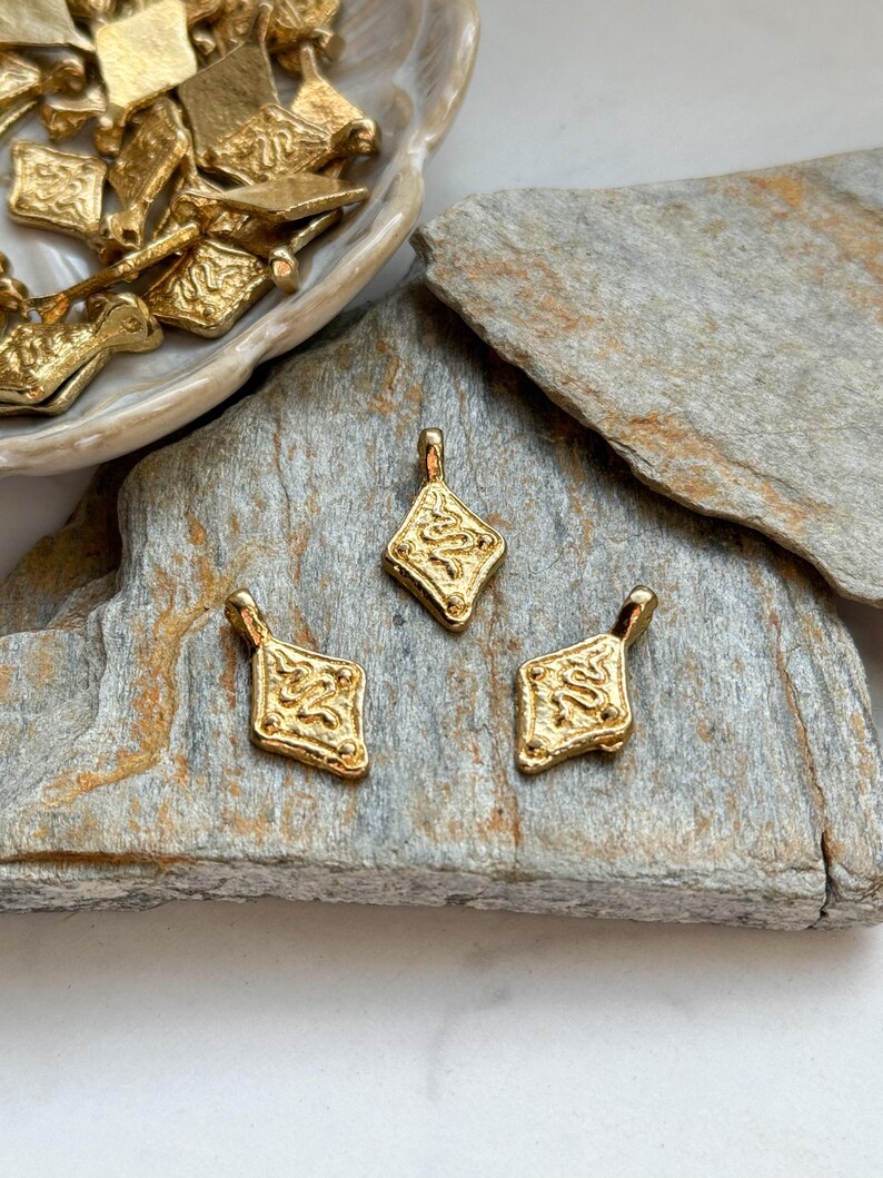 Pode incluir: Pingentes de metal dourado em forma de diamante com design ornamentado. Tr&ecirc;s pingentes s&atilde;o dispostos sobre uma superf&iacute;cie de pedra cinza, com pingentes adicionais em um prato decorativo. Os pingentes t&ecirc;m uma argola na parte superior.