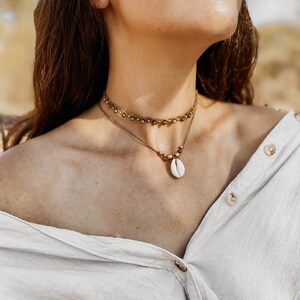 May include: A woman wearing a white button-down shirt with a gold chain necklace featuring a white seashell pendant. The necklace is layered over a gold beaded choker.