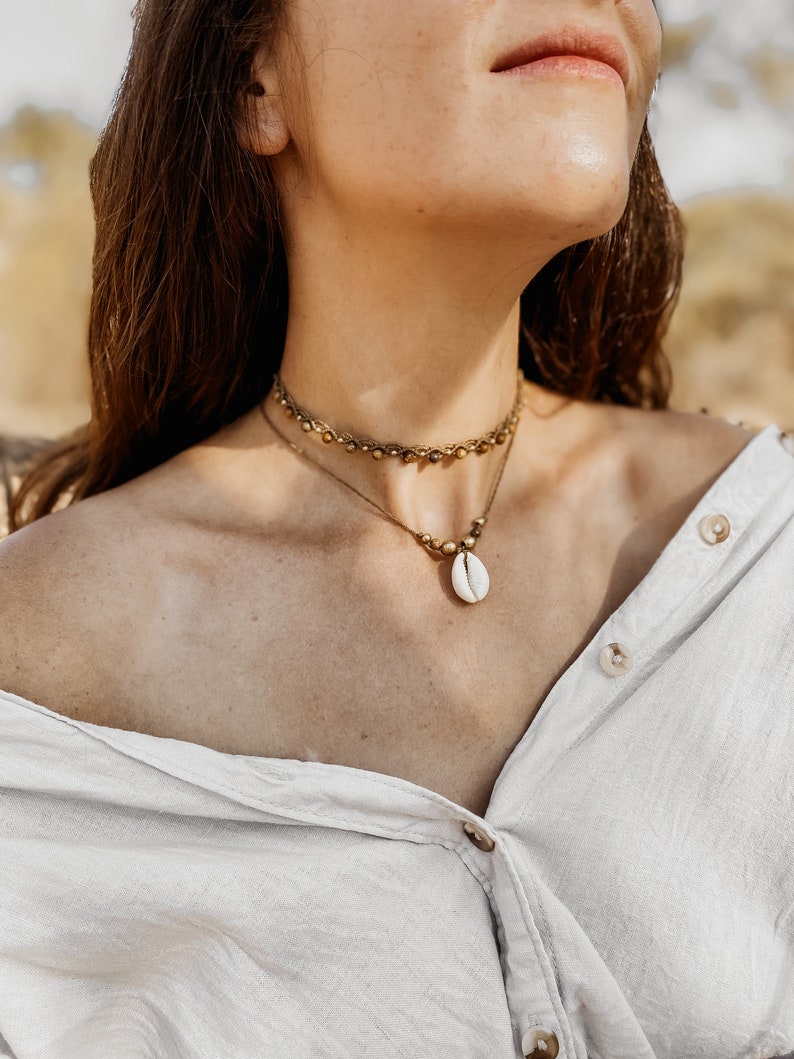 May include: A woman wearing a white button-down shirt with a gold chain necklace featuring a white seashell pendant. The necklace is layered over a gold beaded choker.