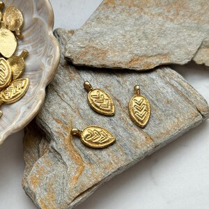 May include: Three gold leaf-shaped charms with a geometric pattern. The charms are on a light brown stone surface.