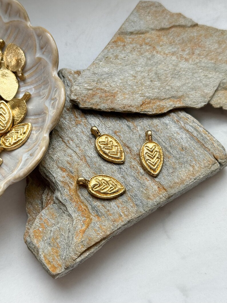 May include: Three gold leaf-shaped charms with a geometric pattern. The charms are on a light brown stone surface.