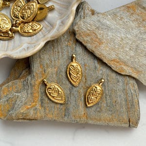 May include: Three gold leaf-shaped charms with a geometric pattern. The charms are on a light brown stone surface.