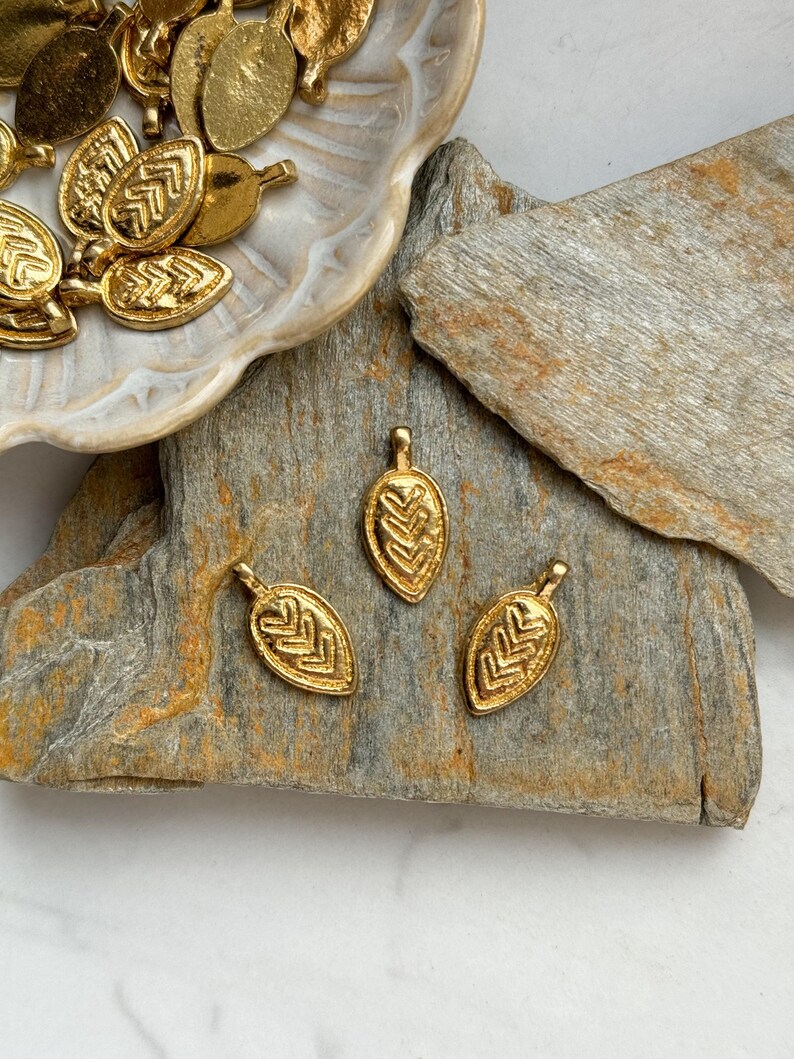 May include: Three gold leaf-shaped charms with a geometric pattern. The charms are on a light brown stone surface.