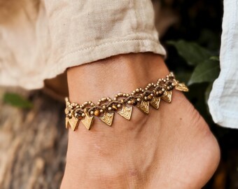 May include: A gold anklet with a braided cord and triangular charms. The anklet is worn on a person's ankle.