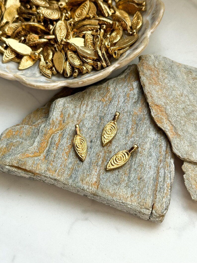 May include: Three gold leaf-shaped charms with a swirl design. The charms have a hole at the top for attaching to jewelry or other crafts.