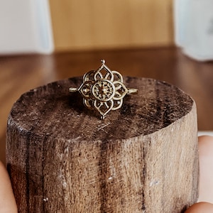 K&ouml;nnte beinhalten: Goldring mit einem filigranen, komplizierten Design, das ein mandalaartiges Muster zeigt. Der Ring liegt auf einer Holzoberfl&auml;che.