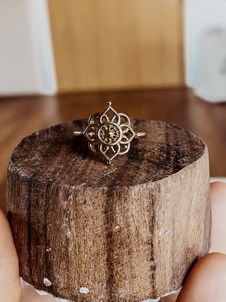 K&ouml;nnte beinhalten: Goldring mit einem filigranen, komplizierten Design, das ein mandalaartiges Muster zeigt. Der Ring liegt auf einer Holzoberfl&auml;che.