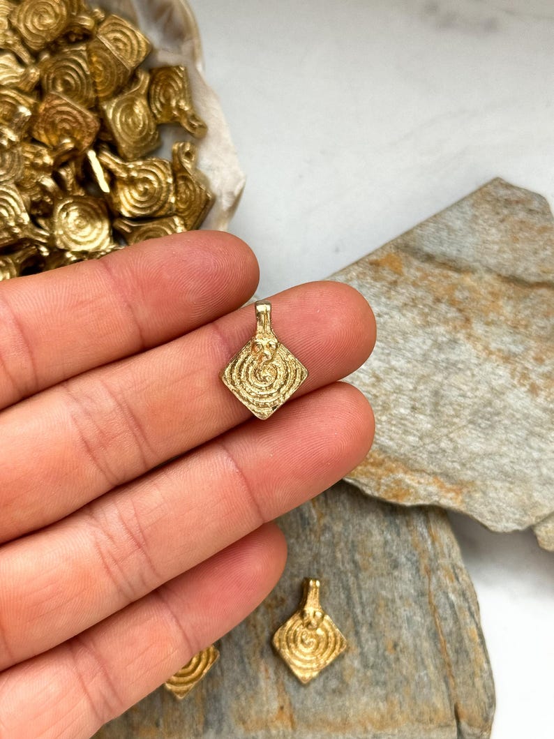 K&ouml;nnte beinhalten: Goldfarbener Metallanh&auml;nger mit quadratischer Form und einem wirbelnden, konzentrischen Kreisdesign. Der Anh&auml;nger hat eine &Ouml;se oben zum Anbringen an einer Kette oder Schnur.