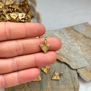May include: A gold-colored metal pendant in the shape of a triangle with a geometric pattern. The pendant is being held in a hand.