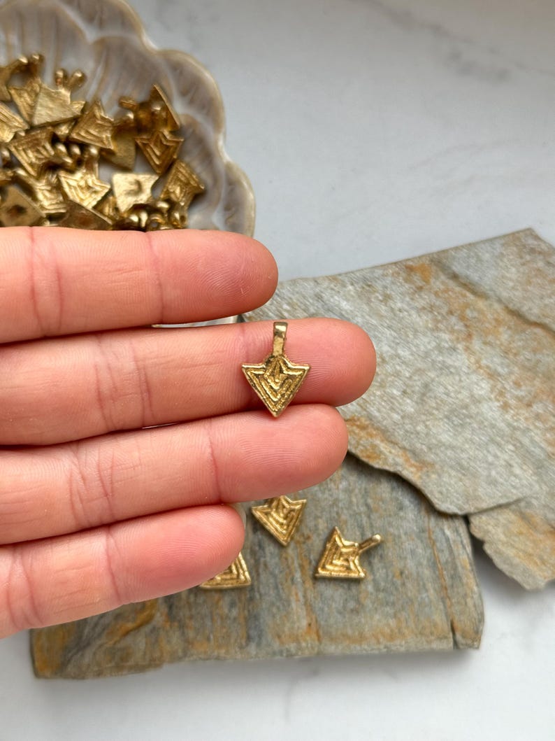 May include: A gold-colored metal pendant in the shape of a triangle with a geometric pattern. The pendant is being held in a hand.