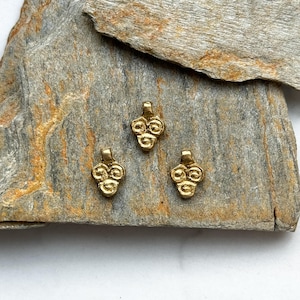 May include: Three gold-colored metal charms in the shape of a cluster of grapes. The charms are on a gray and brown stone surface.