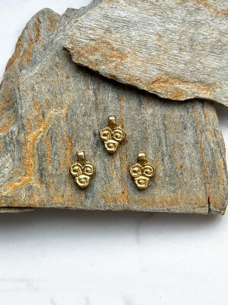May include: Three gold-colored metal charms in the shape of a cluster of grapes. The charms are on a gray and brown stone surface.