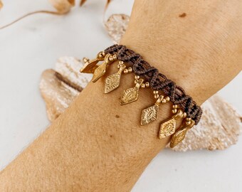 Puede incluir: Un brazalete trenzado marrón con abalorios de metal dorado en forma de diamantes. El brazalete se lleva en la muñeca.