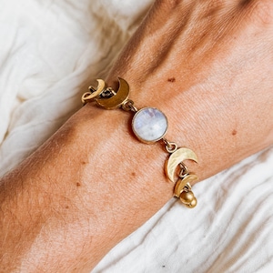 May include: A gold-toned bracelet featuring a central round iridescent moonstone cabochon flanked by multiple crescent moon charms. The metal links connect the moonstone to the crescent shapes, with small gold spheres dangling at the end of the chain.