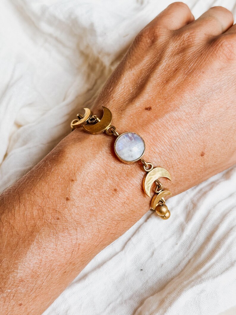 May include: A gold-toned bracelet featuring a central round iridescent moonstone cabochon flanked by multiple crescent moon charms. The metal links connect the moonstone to the crescent shapes, with small gold spheres dangling at the end of the chain.