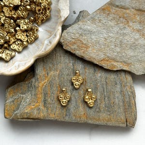 May include: A close-up of three gold-colored metal charms shaped like a cluster of grapes. The charms are sitting on a piece of rough, gray stone.